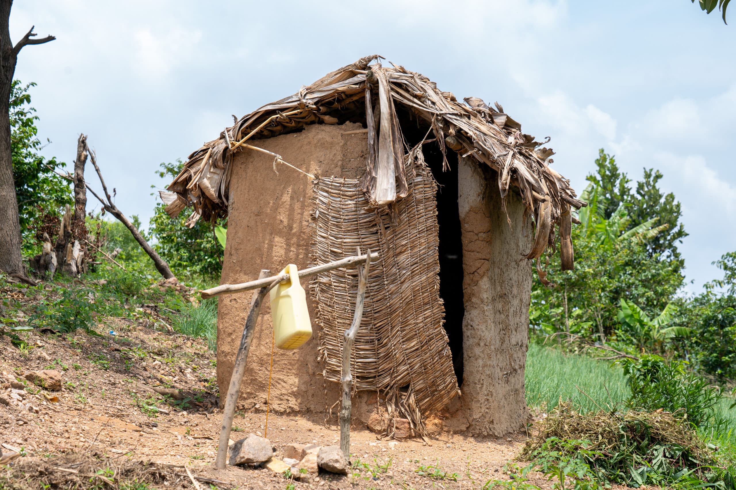 A small, rustic hut, used as a closed pit latrine, made of mud and thatched roof, with a woven mat serving as a door. In front of the hut, a handwashing station with a yellow plastic container is set up, surrounded by natural vegetation in a rural setting