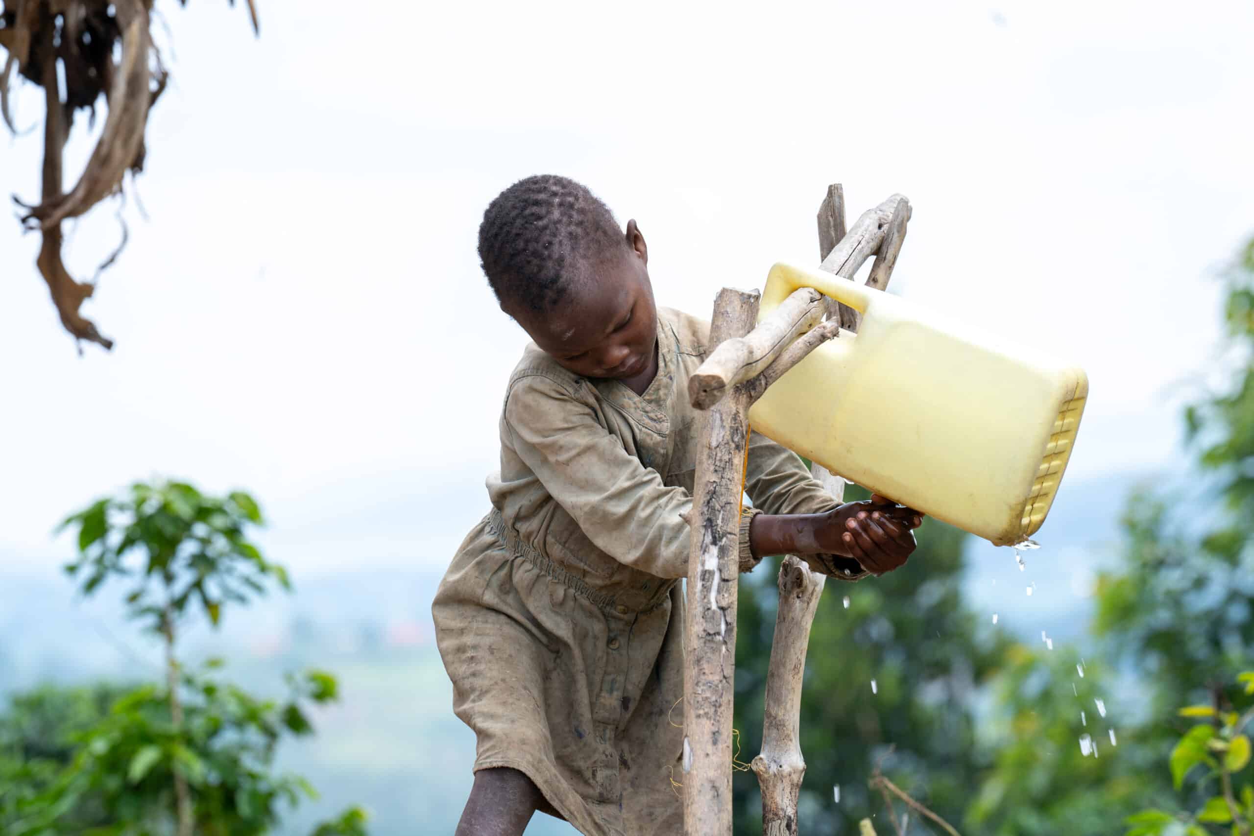 A child washing hands at a handwashing station set up with a yellow plastic container surrounded by natural vegetation in a rural setting."