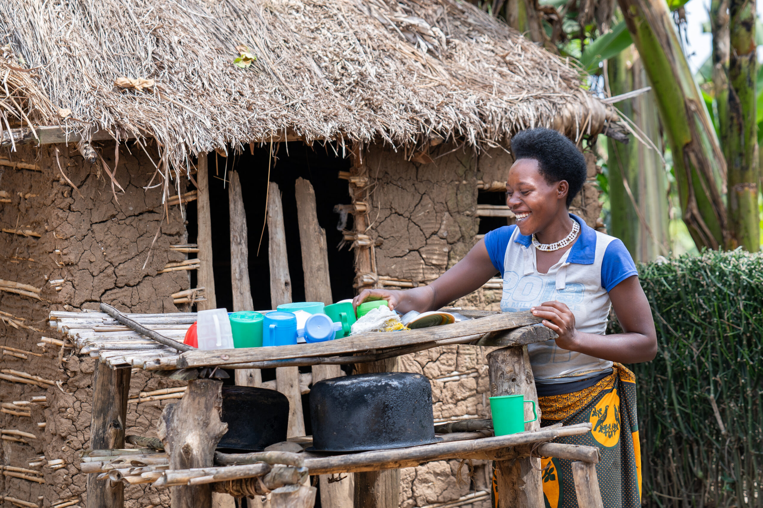 Turinawe using a drying dish rack as a better hygiene practice. A woman smiles as she washes dishes at a wooden dish rack outside a traditional mud and stick house with a thatched roof. She is dressed in a blue-collared T-shirt and a patterned wrap skirt. The dish rack holds several colorful plastic cups and bowls, along with two large black cooking pots. The setting is lush and rural, with green banana plants and hedges in the background.